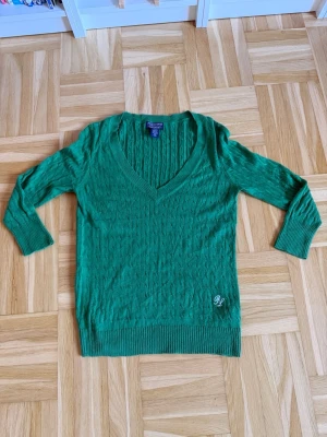 Grön stickad tröja med v-ringning - Grön stickad tröja med djup v-ringning och diskret kabelstickat mönster. Tröjan har långa ärmar, ribbade muddar och en liten broderad detalj med bokstäverna 'RL' nedtill. Perfekt för dig som gillar klassisk men ändå trendig stil.