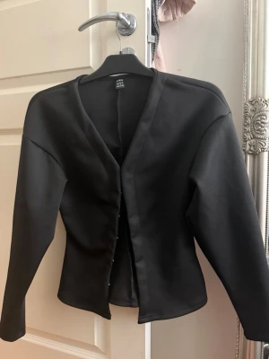 Svart figursydd cardigan - Stilren svart cardigan med v-ringning och knäppning framtill. Figursydd modell med långa ärmar och slät yta. Perfekt att matcha med både jeans och kjol för en trendig look.