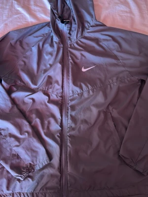 Lila vindjacka från Nike med huva - Snygg lila vindjacka från Nike med dragkedja framtill och huva. Jackan har två sidofickor och den klassiska Nike-loggan på bröstet. Tillverkad i lätt och vindtätt material, perfekt för blåsiga dagar.