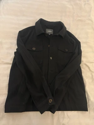 Svart overshirt med fickor - Stilren svart overshirt med klassisk krage och knäppning framtill. Två bröstfickor med lock och knappar samt knappar vid ärmsluten. Materialet ser ut att vara kraftigt och passar perfekt för lager-på-lager. Enkel och clean design som funkar till det mesta.