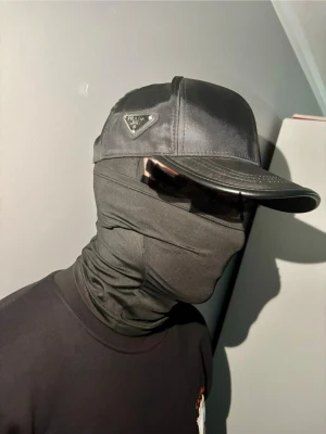 Svart keps från Prada i nylon - Snygg svart keps från Prada i glansigt nylonmaterial med klassisk böjd skärm och ikonisk metallogo på sidan. Kepsen har en stilren och modern look som passar perfekt till streetwear och trendiga outfits.
