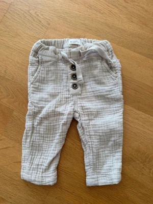 Baby beige linnebyxor med knappar - Bebis beiga linnebyxor i mjukt, luftigt tyg med resår i midjan och dekorativa knappar framtill. Byxorna har sidfickor, bakfickor och ett knytband i midjan för extra stilpoäng. 
