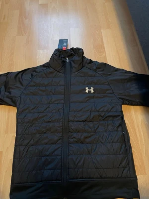 Under armour jacka  - This is a men's black Under Armour puffer jacket. The front features a quilted design with horizontal stitching and a full zip closure. The Under Armour logo is visible on the left chest in a contrasting color. The back of the jacket has reflective horizontal bars near the collar for visibility. The jacket has an athletic fit and is designed for warmth and comfort, suitable for casual or athletic wear.