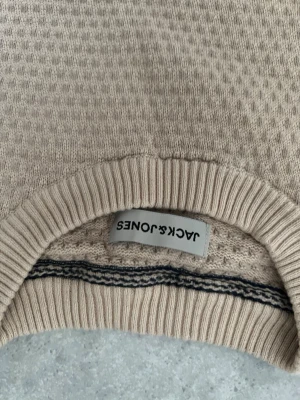 Beige stickad tröja från Jack & Jones - Snygg beige stickad tröja från Jack & Jones med rund halsringning och ribbade muddar. Tröjan har ett diskret mönster i stickningen och passar perfekt till en avslappnad stil. Materialet känns mjukt och bekvämt.