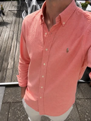 Polo Ralph Lauren Skjorta – Rosa Slim Fit Oxford - Snygg Polo Ralph Lauren skjorta i en stilren rosa/coral färg. Modellen är Slim Fit Stretch Oxford och har klassisk button-down-krage samt det ikoniska broderade Polo-ponnyloggan på bröstet.  • Storlek M • Slim Fit • Stretch Oxford • Rosa/coral • Button-down krage • Polo Ralph Lauren.                                       Skjortan är i fint skick och passar perfekt till både jeans, chinos eller shorts. En riktigt klassisk Ralph Lauren-skjorta som fungerar både till vardags och lite mer uppklätt.