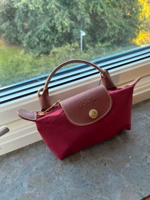 Vinröd  handväska - Snygg vinröd handväska från  med bruna detaljer i läder och guldfärgade metalldetaljer. Väskan har ett klassiskt veckat lock med tryckknapp och dragkedja. Perfekt storlek för det viktigaste och enkel att bära i handtaget.
