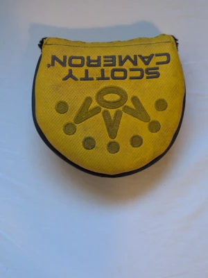 Scotty Cameron headcover - Säljer ett gult Scotty Cameron headcover till golfklubba. Fodralet har broderad text och mönster i svart och brunt samt en unik design med rundad form och mjukt material. Perfekt för dig som vill skydda din putter med stil.