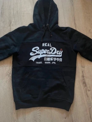 Svart hoodie från Superdry - Snygg svart hoodie från Superdry med stor logga och japansk text på bröstet. Hoodien har huva med dragsko och en stor magficka. Materialet ser mjukt och bekvämt ut, perfekt för en avslappnad stil. Diskret camomönster på nedre delen ger extra edge.