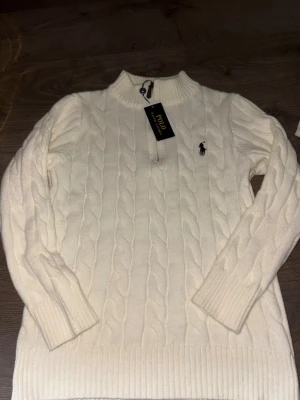 Vit kabelstickad tröja Polo Ralph Lauren - Halv zip kabelstickad vit tröja från Polo Ralph Lauren med rund halsringning och klassisk logga broderad på bröstet. Tröjan har långa ärmar och ribbade muddar vid ärmslut och nederkant. Perfekt för en stilren och tidlös look.