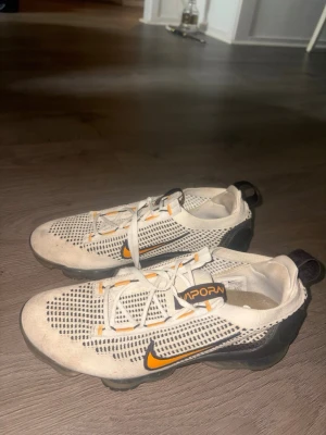 Nike Air VaporMax vita sneakers - Säljer mina vita Nike Air VaporMax. Knappt använda och i väldigt fint skick. Köpta för 2 500 kr. Nike Air VaporMax sneakers i vitt med svarta och orange detaljer. Ovandelen är i mesh för bra ventilation och skon har den ikoniska genomskinliga sulan med synliga luftkuddar. Snygg och sportig design med snörning och dragflik bak. 