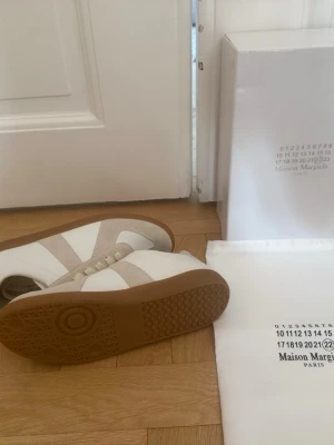 Maison Margiela vita sneakers - Säljer ett par vita sneakers från Maison Margiela med beige detaljer och gummisula. Skorna har en klassisk låg silhuett och är tillverkade i en mix av läder och mocka. Perfekta för dig som gillar stilrena och exklusiva sneakers.