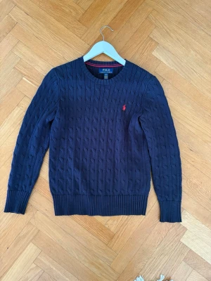 Mörkblå kabelstickad tröja Polo Ralph Lauren - Snygg mörkblå kabelstickad tröja från Polo Ralph Lauren med rund halsringning och röd broderad logga på bröstet. Tröjan har ribbade muddar vid ärmslut och nederkant. Perfekt för dig som gillar klassisk och stilren design.