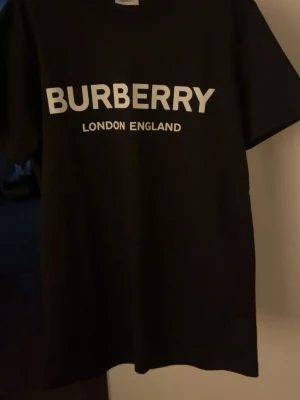 Svart Burberry t-shirt med tryck - Säljer en svart t-shirt från Burberry med stort vitt tryck på bröstet där det står 'BURBERRY LONDON ENGLAND'. Klassisk passform och rund hals. Perfekt för dig som gillar stilrena och ikoniska plagg.