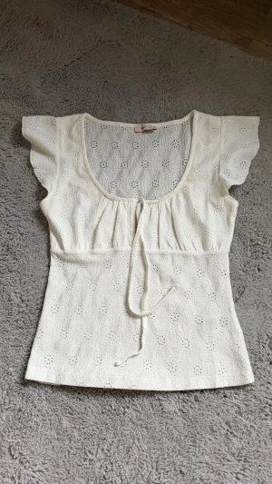 Vit broderad blus med volangärm - Söt vit blus med broderade detaljer och volangärmar. Modellen har en rundad urringning med knytband framtill och är tillverkad i ett lätt bomullstyg. Perfekt för en romantisk och somrig look.