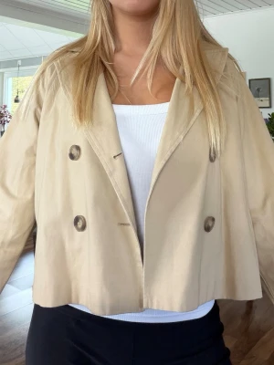 Kort beige trenchcoat med dubbelknäppning - Skitsnygg kort beige trenchcoat. Passar perfekt till vår och höst!