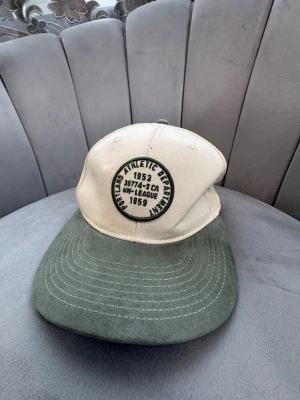Beige och grön keps med brodyr - Snygg keps i beige med grön skärm och broderad patch framtill där det står 'Portland Athletic Department 1953'. Kepsen har klassisk form och justerbar baksida. Materialet ser ut att vara bomullstyg, perfekt för en avslappnad streetwear-look.