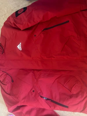Röd vindjacka från Adidas med huva - Snygg röd vindjacka från Adidas med dragkedja, huva och flera praktiska fickor med dragkedja. Jackan har Adidas-logga på bröstet och en patch på ärmen. Perfekt för blåsiga dagar och sportiga outfits.