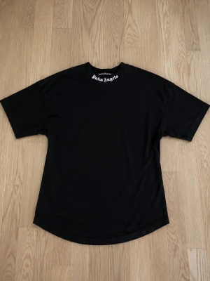 Svart Palm Angels t-shirt - Säljer en svart t-shirt från Palm Angels med logga tryckt i vitt på både framsidan vid halsen och stort på ryggen. T-shirten har rund hals, korta ärmar och är gjord i mjuk bomull. Perfekt för dig som gillar streetwear och vill sticka ut.