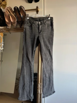 Grå slitna bootcut jeans - Säljer ett par grå bootcut jeans med slitningar på ena benet. Jeansen har klassisk femficksdesign och är tillverkade i ett mjukt denimtyg med lätt stretch. Perfekta för en avslappnad och trendig look.