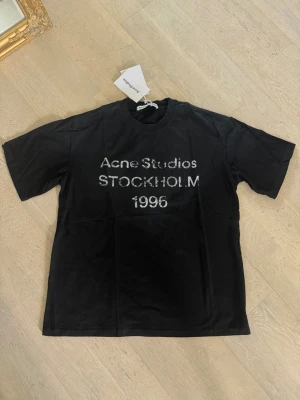 Svart Acne Studios t-shirt - Säljer en svart t-shirt från Acne Studios med trycket 'Acne Studios STOCKHOLM 1996' i vitt framtill. Klassisk rund hals och korta ärmar. Perfekt för dig som gillar stilrena och trendiga plagg med streetkänsla.