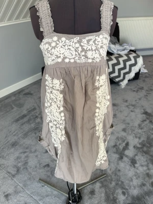 Beige klänning med spets från Superdry - Säljer en beige kortklänning från Superdry med breda axelband och vackra vita spetsdetaljer och broderier framtill. Klänningen har en avslappnad passform och är perfekt för varma dagar. Märkt med Superdry Vintage Thrift.