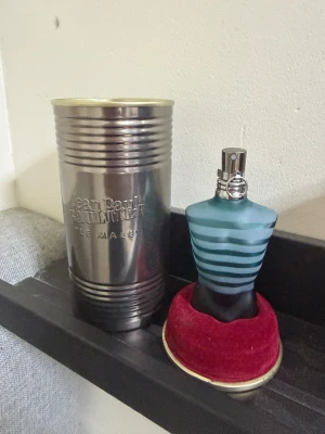 Jean Paul Gaultier Le Male parfym - Klassisk parfymflaska från Jean Paul Gaultier i form av en manlig torso med blå och turkosa ränder. Flaskan står i en röd sammetsbas och kommer med en metallisk cylinderformad förpackning. En ikonisk doft som sticker ut i badrumshyllan. Har ungefär halva i sig 