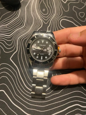 Klassisk silverfärgad armbandsklocka - Stilren armbandsklocka med svart urtavla och silverfärgad länk i metall. Klockan har tydliga siffror och markeringar på boetten samt en orange detalj på sidan. Perfekt accessoar för en modern look.