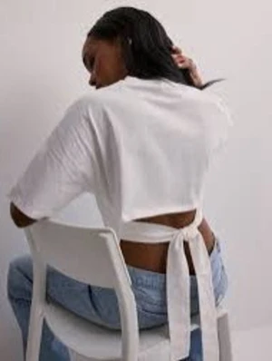 Vit crop top med knytband bak - Trendig vit crop top med korta ärmar och breda knytband i ryggen som ger en öppen ryggdetalj. Toppen har en boxy passform och är gjord i ett mjukt, tunt tyg som känns skönt mot huden. Perfekt att styla med höga jeans eller kjol.