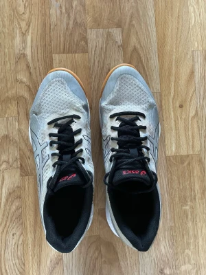 Asics silvriga inomhusskor - Säljer ett par silvriga Asics inomhusskor med svart snörning och detaljer. Skorna har svart sula med gummifärgad undersida och röda inslag vid hälen. Perfekta för sporter inomhus. Storlek 42.5 Eu. Har också 41.5 om 42.5 är för stort