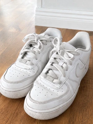 Vita Nike Air Force 1 sneakers - Klassiska helvita Nike Air Force 1 sneakers i lågt utförande. Skorna har en stilren design med perforerade detaljer på tån, kraftig sula och Nike-logga på sidan och hälen. Tillverkade i läder med vita skosnören och bekväm vadderad insida.