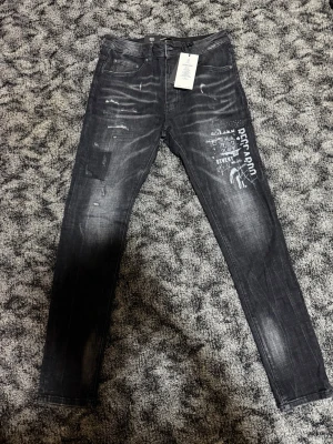 Svarta jeans med tryck från Reccardo Sevent - Snygga svarta jeans från Reccardo Sevent med slitningar, patchdetaljer och vitt tryck på ena benet. Jeansen har en smal passform och klassisk femficksdesign. Materialet är denim och designen är italiensk. Perfekta för en trendig streetwear-look.