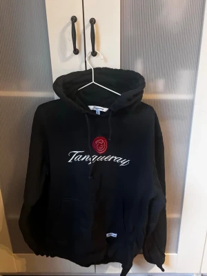 Svart Tanqueray hoodie - Svart hoodie från Tanqueray med vit broderad logga och röd detalj framtill. Klassisk känguruficka och justerbar huva. Perfekt för dig som gillar streetwear och vill ha en chill look. Mjuk bomullskänsla och avslappnad passform. Säljes då den ej passar de är Storlek L men sitter mer som M