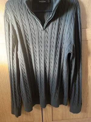 Mörkgrå kabelstickad tröja med halvzip - Snygg mörkgrå kabelstickad tröja från Dressmann med halv dragkedja i halsen. Tröjan har långa ärmar, ribbade muddar och klassiskt kabelmönster över hela plagget. Perfekt för dig som gillar en stilren och tidlös look.