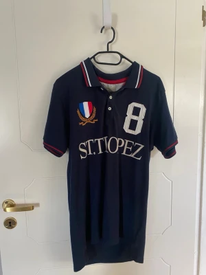 Marinblå pikétröja St. Tropez - Marinblå pikétröja från Selected Homme med broderad St. Tropez-text, stort vitt nummer 8 och fransk sköld på bröstet. Kragen har röda och vita ränder och tröjan har två knappar framtill. Klassisk och sportig stil.