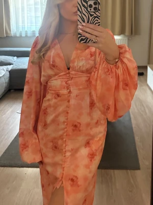 Adoore klänning positano - Romantisk långklänning i ljusa rosa och orange toner med blommigt mönster. Klänningen har v-ringning, puffiga långärmar och knappar framtill. Tillverkad i ett lätt, flytande material som ger en elegant siluett.