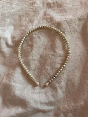 Pärldiadem i vitt - Snyggt diadem dekorerat med vita pärlor längs hela bågen. Klassisk och stilren design som ger en elegant touch till din look. Diademet har rundad form och pärlorna ger en lyxig känsla. Perfekt accessoar för att lyfta vilken outfit som helst. Pärlorna är fästa på en stomme av metall som har rostat något i nedre kanten (se bild).