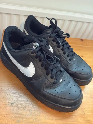 Nike Air Force 1 svarta sneakers - Klassiska Nike Air Force 1 sneakers i svart läder med vit swoosh och AIR-logga på hälen. Skorna är i fint skick och har en clean look. Perfekta till vardags eller för en snygg streetwear-outfit.Skorna är använda.                 
