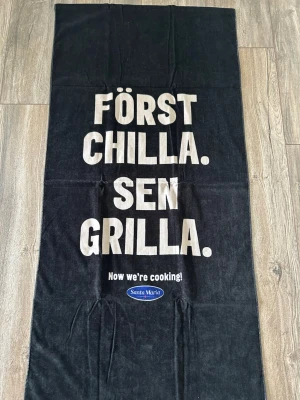 Svart strandhandduk Santa Maria - Stor svart strandhandduk med vit text 'FÖRST CHILLA. SEN GRILLA.' och Santa Maria-logga. Perfekt för stranden eller picknicken, tillverkad i mjukt material och har en cool design med tydligt budskap.
