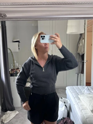 Mörkgrå cropped hoodie med dragkedja - Säljer en mörkgrå cropped hoodie med dragkedja framtill och ribbade muddar. Hoodien har en huva med dragsko och är tillverkad i ett stickat material som ger en avslappnad och trendig look. Perfekt till jeans eller shorts för en chill stil.