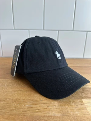Svart keps från Polo Ralph Lauren - Snygg svart keps från Polo Ralph Lauren med klassisk böjd skärm och vit broderad logga framtill. Tillverkad i bomullstyg och har justerbar rem baktill för perfekt passform. Perfekt accessoar för en clean och stilren look.