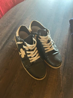 Svarta sneakers med vit detalj - Säljer ett par svarta sneakers med vita skosnören och en stor vit klöverdetalj på sidan. Skorna har vit sula och vit hälkappa. Klassisk låg modell som passar till det mesta. Materialet ser ut att vara läder eller konstläder.