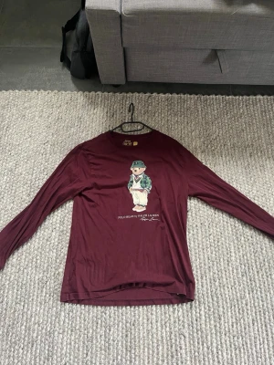 Vinröd långärmad Polo Bear tröja - Säljer en vinröd långärmad tröja från Ralph Lauren med det ikoniska Polo Bear-motivet på bröstet. Tröjan har rund hals och är gjord i mjukt material, perfekt för en avslappnad och stilren look. ÄR I BARN XL, passar vanlig medium