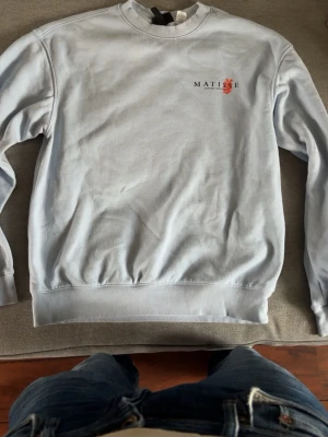 Ljusgrå sweatshirt med Matisse-tryck - Säljer en ljusgrå sweatshirt med rund hals och ribbade muddar. På bröstet finns ett litet Matisse-tryck och på ryggen ett stort orange konstmotiv samt text. Perfekt för dig som gillar konst och streetwear-vibe.