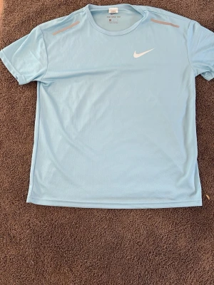 Ljusblå Nike sport t-shirt - Snygg ljusblå sport t-shirt från Nike med rund halsringning och vit logga på bröstet. Tillverkad i ett lätt och ventilerande material som passar perfekt till träning eller löpning. Klassisk passform och korta ärmar.