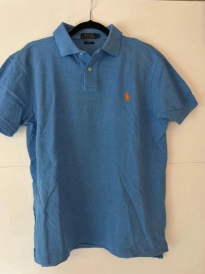Blå pikétröja från Polo Ralph Lauren - Snygg blå pikétröja från Polo Ralph Lauren med klassisk krage, korta ärmar och orange logga på bröstet. Slim fit-modell i mjuk bomull som passar perfekt till jeans eller shorts. Två knappar vid halsen för justerbar stil.