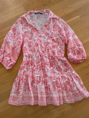 Rosa blommig kortklänning med volang - Supersöt kortklänning i rosa och vitt med blommigt mönster och volangkant. Klänningen har trekvartsärmar, markerad midja och en avslappnad passform. Perfekt för varma dagar och somriga vibes!