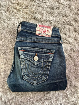 True Religion lowwaist blå jeans med kontrastsöm - Säljer ett par blå lowwaist jeans från True Religion med ikoniska kontrastsömmar och stor ficka bak med knapp. Klassisk patch i vitt och rött bak i midjan. Snygg tvättad look och smal passform. Perfekta till en avslappnad streetstyle.
