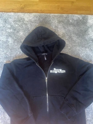 Svart hoodie med tryck och dragkedja - Svart hoodie med dragkedja framtill och vit texttryck på bröstet där det står 'Avenue des Champs-Élysées Montaigne Georges V'. Hoodien har huva och kängurufickor. Mjuk insida och klassisk passform, perfekt för en chill och avslappnad stil.