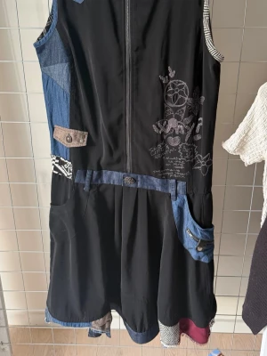 Svart ärmlös klänning med denimdetaljer - Unik svart klänning med dragkedja framtill, denimdetaljer på fickor och midja samt broderade mönster på bröstet. Flera fickor och kontrasterande tygdetaljer i blått och beige ger en cool patchwork-känsla. Perfekt för dig som gillar en kreativ och avslappnad stil.