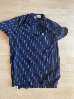 Marinblå randig t-shirt från FILA - Snygg marinblå t-shirt från FILA med vita vertikala ränder och liten logga på bröstet. Klassisk rund hals och korta ärmar. Perfekt för en sportig och avslappnad stil.